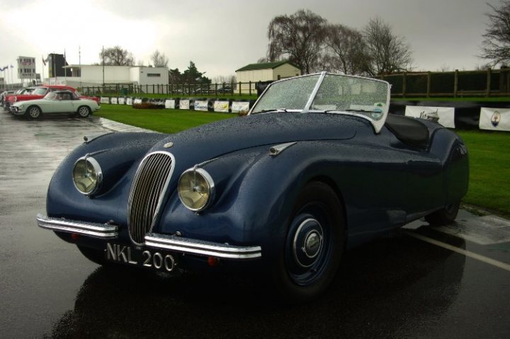 Pistonheads - The image captures a scene on a rainy day in front of a building with a vintage blue sports car as the focal point. The car bears the registration NKL 200 and is a classic model resembling a Jaguar or similar British sports car commonly seen in race events in the early 1900s. The car's metallic blue exterior gleams under the cloudy sky, its headlights and tail lights clearly visible. In the background, there is a small white building and several other cars, indicating a public area or event. The wet road reflects the car and the surroundings, adding a reflective quality to the image.