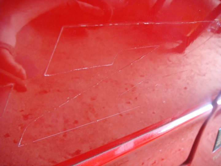 Removed Pistonheads Laquered Decals - The image shows a close-up view of a red car with a glossy finish. There are two prominent marks on the red vehicle that appear to be key scratches. One mark is in the middle, and the other is towards the right side. The reflection of the car in the glass nearby highlights the shiny surface and the scratches. The surface appears to be wet, giving the reflection a slightly distorted effect. The overall condition of the car suggests that it has been used, and there might have been a previous incident involving keys.