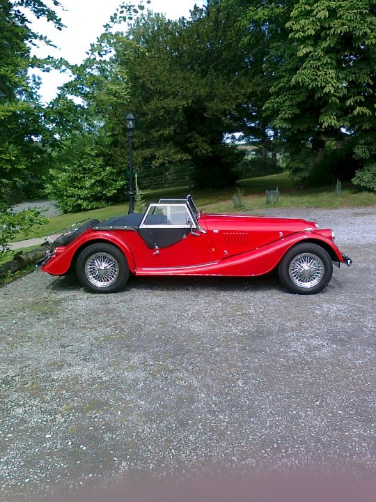 REALLY got a thing for a Morgan now - Page 1 - Morgan - PistonHeads - The image shows a vintage sports car parked on a gravel surface. The car is predominantly red, with a black roof and contrasting silver accents around the windows and on the body. It has a sleek, aerodynamic profile with a distinct long hood and short rear deck. The vehicle features spoked wheels and a large front grille with a badge, which is difficult to read. The scene includes trees in the background, suggesting a natural environment. The lighting in the image is bright and even, indicating common daylight conditions.