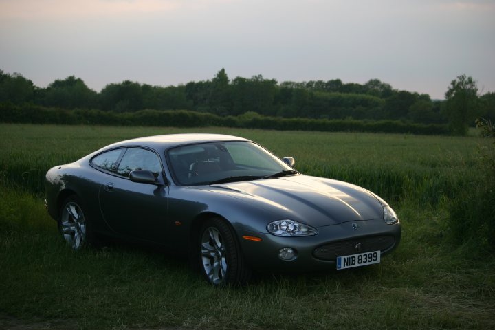 A black and white photo of an old car - Pistonheads - The image shows a black sports car parked in a grassy field. The car has a sleek and streamlined design, typical of sports vehicles. It is facing towards the left side of the photo, and the setting sun casts a warm glow on the car. The background features a large open field, hinting at a rural or rural-fringe location. The sky is overcast, suggesting that this scene might be captured in late afternoon or early evening.