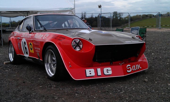 any 240z owners here? - Page 1 - Jap Chat - PistonHeads - The image showcases a vibrant red and white sports car on a gravel track. The car is adorned with the name "Big Sam" on its bumper. Behind the car, two people can be seen, likely the car's owners or mechanics. A fence lines the back of the field, suggesting that this could be an open track or race circuit. The sky overhead is partly cloudy, casting a soft light on the scene.