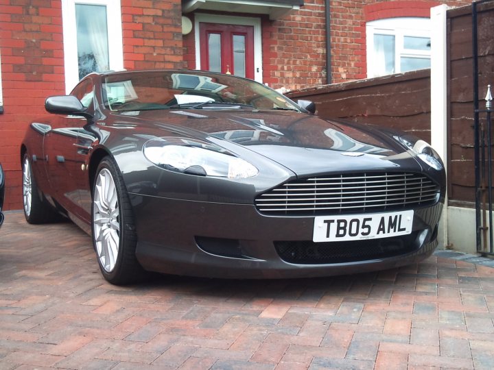 Have to tell someone - Page 1 - Aston Martin - PistonHeads - The image shows a sleek, dark-colored sports car parked on a brick driveway in front of a house. The car is a convertible with a distinctive design and wheel rims. At the front of the car, there is a license plate that reads "TBO5 AML." The vehicle's condition appears to be well-maintained, and it stands out against the backdrop of the house. The overall setting suggests it might be a residential area.