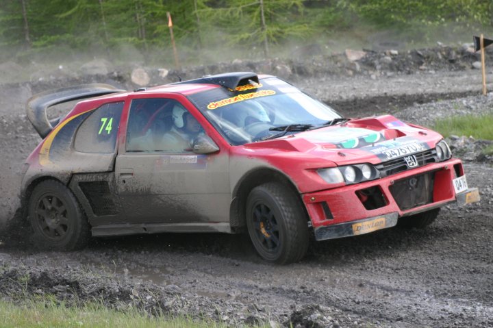 Pistonheads Sunseeker Rally - The image showcases a thrilling moment on a muddy race track. A red rally car, #74, is captured mid-action, kicking up a cloud of mud as it navigates the course. The battered vehicle, crushed and dented from use, is adorned with a label reading "001". It's clear that this is an event filled with adrenaline and action. The driver and co-driver, hidden from view, are immersed in the world of rally driving.