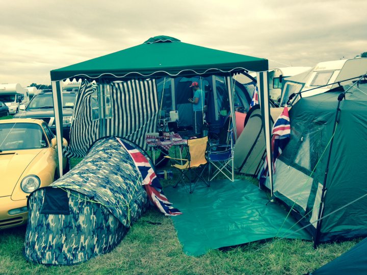 A group of lawn chairs sitting next to each other - Pistonheads - The image depicts a campsite setting, with several tents pitched on green grass. A prominent umbrella-shaped tent stands in the foreground, while various other tents of varying shapes and sizes are scattered around. Two cars, a yellow and a black vehicle, are parked nearby, and there is a dining table and chairs within the immediate vicinity of the image, surrounded by camping equipment. The scene captures a warm and casual outdoor gathering or camping trip.