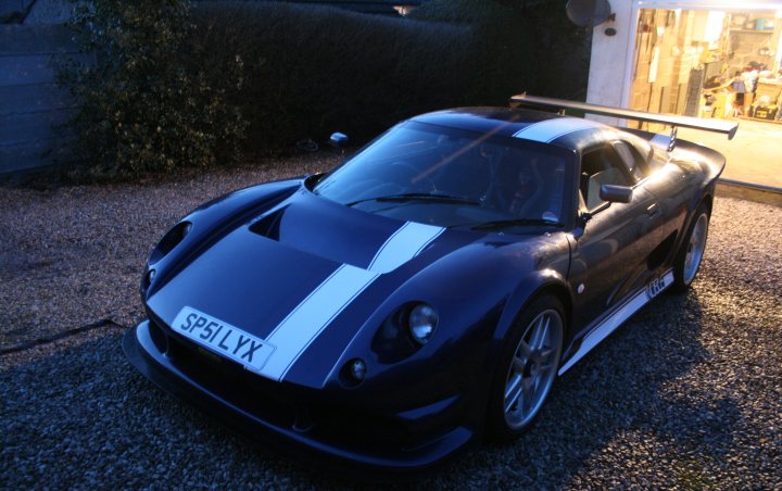 Introduction Pistonheads Photoshoot Free - The image depicts a striking black and white sports car, parked on gravel. The car boasts a sleek design with a prominent front bumper and large wheels that hint at its performance capabilities. The setting sun casts a dramatic light on the car, enhancing its sporty silhouette against the darker blue sky. The background features an outdoor area illuminated by artificial light, softly blurred to focus attention on the car in the foreground.