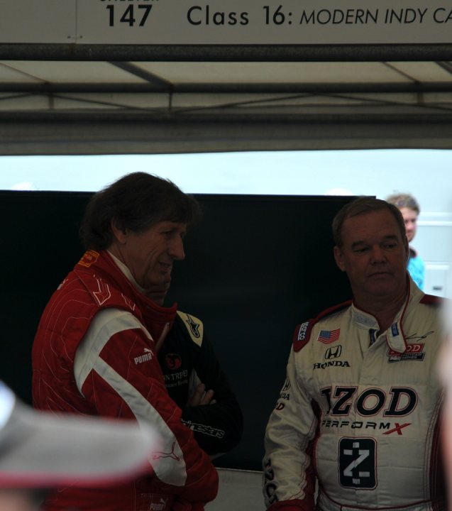 Post your Pics, Celebrities at  FOS 2011 - Page 1 - Goodwood Events - PistonHeads - The image showcases two racing drivers at a race track, possibly between sessions or before a race. They are in conversation, one standing and the other slightly leaning on a black wall. Both drivers are wearing red racing suits, indicative of their teams. The driver on the right side of the image is looking directly at the camera, while the one on the left appears to be looking away, possibly focusing on something in his peripheral vision. The drivers' expressions convey a serious yet competitive atmosphere typical of the sport.