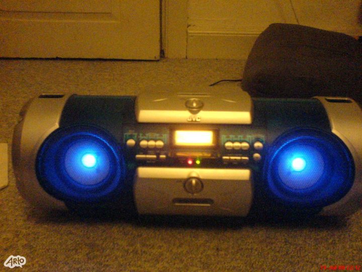 A remote control sitting on top of a table - Pistonheads - The image showcases a high-tech boombox wrapped in blue lights. With a prominent black and gray body, the boombox is placed on a gray carpeted floor. A glimpse of white paint can be seen in the background, creating a contrasting backdrop for the gleaming appliance. Here's four sentences:

1. The boombox, a fusion of intricate electronics, sits in the foreground of the image.
2. It's blanketed in blue lights that give it a modern, futuristic look.
3. The background features a glimpse of white paint on a door, hinting at a homey, contrasting backdrop.
4. The gray carpeted floor provides a neutral base that allows the blue-lighted boombox to stand out.