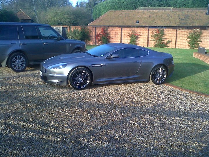 Pistonheads - The image showcases a luxury sports car parked on a gravel surface. The car has a sleek design with a glossy paint finish reflecting the sunlight. In the background, there is a gray SUV and a brick wall, adding a contrasting to the modern appearance of the sports car. The bright daylight illuminates the scene, highlighting the refined details of the car's design.