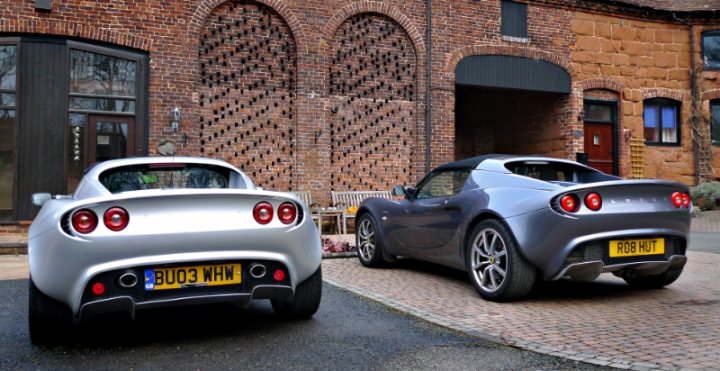 Headlamps on an 111R '06. - Page 1 - Elise/Exige/Europa/340R - PistonHeads - The image is a daylight photograph of two cars parked in front of a building, likely a parking lot. One car is silver, and the other has a body kit in a similar color palette, possibly the same vehicle with an aftermarket enhancement. The background features a building with a brick facade, featuring arched windows and a solid door. There are no visible people or distinct movement in the image, suggesting a still, perhaps a morning or afternoon time. The overall style of the image is realistic, typical of automotive photography meant to showcase vehicles.