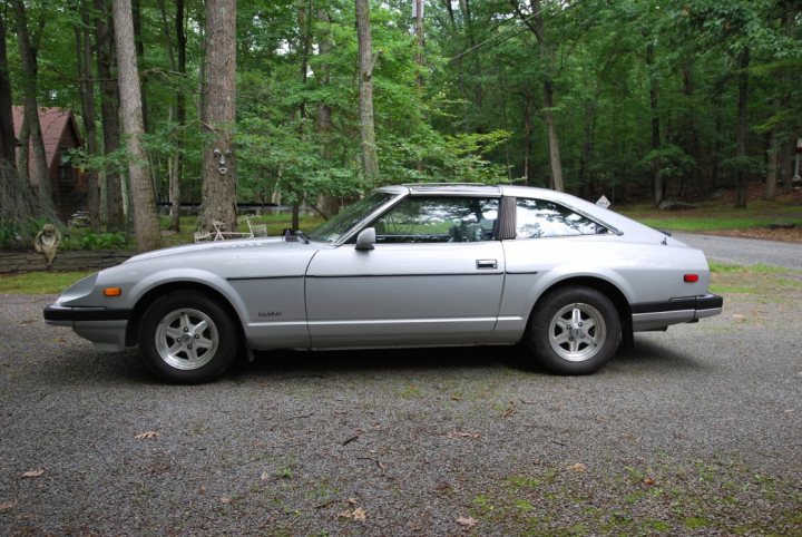 Need advice on a 280ZX can anyone help? - Page 1 - Jap Chat - PistonHeads - The image features a silver 1980s-era sports car parked in a gravel driveway surrounded by trees. The car has a low and sleek profile, with a long hood and a curved rear end, characteristic of classic sports cars. Its design suggests it's a vintage coupe, possibly a Lexus or Toyota with a fair amount of customization. It has a set of large aftermarket wheels and a subtle aftermarket spoiler. The car is positioned facing slightly to the left of the frame, and the environment is serene with a clear view of the woods beyond the gravel drive.