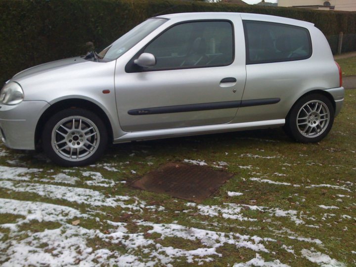 Pistonheads - The image portrays a silver hatchback vehicle parked on a grassy area, covered with a light dusting of snow. The car is positioned at an angle to the camera, allowing a clear view of its front and side frames. The vehicle features alloy wheels and a side mirrow. In the background, the outlines of hedges suggest a boundary or border of the parking area. The overall setting seems quiet and peaceful with a soft cover of snow indicating a cold, possibly winter day.