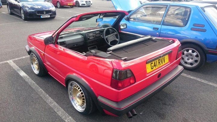 Pictures of decently Modified cars [Vol. 2] - Page 220 - General Gassing - PistonHeads - The image displays a vibrant red convertible car positioned in a parking lot. The car is parked next to a blue hatchback vehicle, indicating that the photo captures a multi-car scene, likely a public parking area. The convertible top is up on the red car, and the wheel is turned towards us, offering a clear view of the car's design. There are no texts or distinctive branding visible in the image. The parking lot appears to have multiple spaces and vehicles, suggesting that this might be a typical urban parking setting.