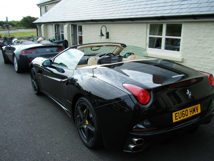 Breakfast Meeting, The Hare, Roxwell 4th June - Page 1 - Kent & Essex - PistonHeads - The image showcases a Rosso Giorgio Concorso spyder parked in front of a white house. A yellow license plate is prominently displayed on the black sports car. In the background, another car is parked, adding to the scenic view.