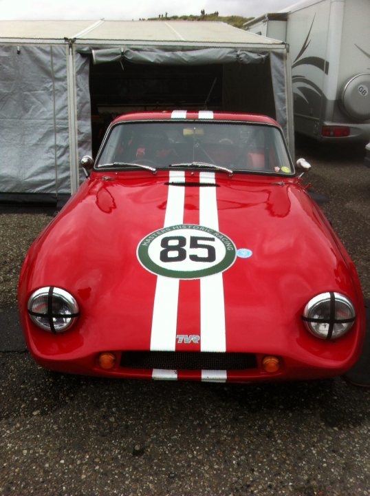 Early TVR Pictures - Page 10 - Classics - PistonHeads - The image displays a classic sports car with a vibrant combination of red and white colors. It features a number "85" prominently displayed on the front bonnet area, suggesting its involvement in racing or a sports-oriented event. The car is parked outdoors, possibly in a temporary parking area, and it is viewed from the front side, with the hood partially open. In the background, there is a tent and part of a trailer, indicating an outdoor event or possibly a show where such classic vehicles are likely to be displayed.