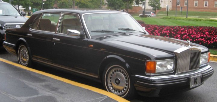 A black and white photo of a car parked in a parking lot - Pistonheads - The image depicts a small, dark-colored, antique sedan parked by the side of a street. Its bright yellow curb adds a vibrant contrast to the car's color scheme. The car is parked on a sidewalk next to a neatly maintained flower bed. In the background, there are trees and buildings, suggesting an urban area. Beyond the parking area, there are two cars parked further away, visible through the car's side window.