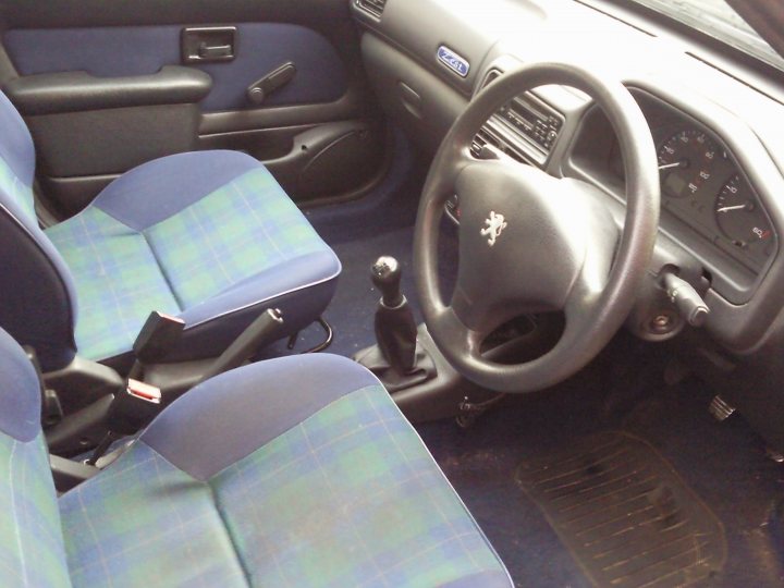 My Beetle and 106 - Page 1 - Readers' Cars - PistonHeads - This image shows the interior of a car at what appears to be a tilted angle, suggesting the photo may have been taken during a car show or at an outdoor event. The seats are upholstered in a blue plaid pattern, with the front seat featuring a gray headrest and a black top. The steering wheel, which is on the left side, has a silver crest on it, indicating it's from a specific brand of car. The dashboard is visible and includes various gauges and controls. In the footwell area, there is a metal floor cover fitted, providing protection for the carpet beneath.