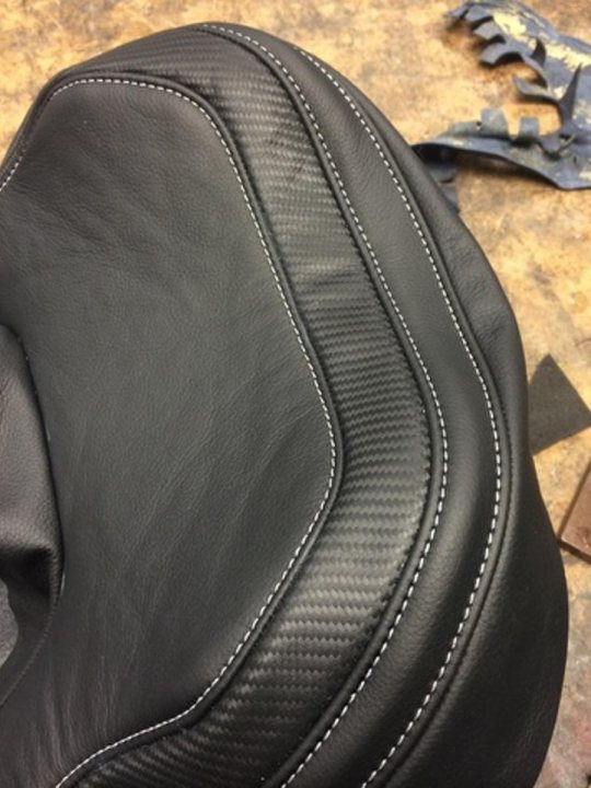 A pair of shoes sitting on top of a suitcase - Pistonheads - The image shows a close-up of a black leather item, possibly a handbag or a piece of motorcycle gear. The architecture of the leather features a curved front panel with stitched lines highlighting the decorative elements. The texture of the leather is a mix of matte and glossy aspects, suggesting high-quality material and craftsmanship. The background appears to be a workspace, with a few scattered items indicating a working environment.