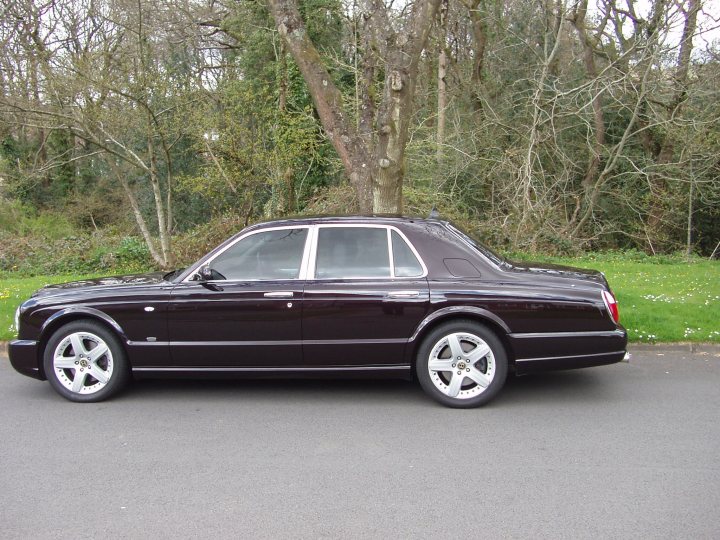 A black and white photo of a car parked in a parking lot - Pistonheads - The image shows a parked dark-colored, possibly black, vintage car. It has four doors, including both front doors and rear carriage-style coach doors. The car is placed on a road with lush greenery in the background, and the setting suggests a calm, possibly suburban, environment. The tire is mounted on a chrome hubcap, indicative of the vehicle's elegant and classic design.