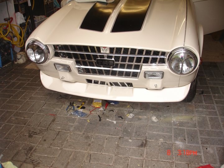 TR6 - Page 1 - Triumph - PistonHeads - This image shows the front view of an old, classic white car with a long hood. The car is facing towards the left side of the photo. It appears to be parked on a tiled floor, which is bathed in a soft yellowish-light, likely due to the natural light coming from the left side of the image. There are repair tools scattered around the periphery of the vehicle, suggesting that maintenance or restoration work might be underway or recently completed. A water mark is visible in the bottom right corner of the image with the numbers "8 3:10 PM" indicating the time the photo was taken or the duration of the exposure.