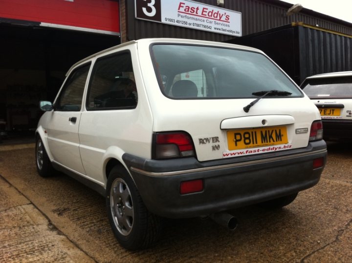 New Car Sneak Preview - Page 1 - East Anglia - PistonHeads - The image shows the rear of a white hatchback parked on a concrete surface. The car features a yellow license plate that reads "P81 MKW." A tribute or advertisement sticker with the text "ROVER IR80" is visible on the back spoiler. In the background, there are industrial structures including a garage with an open door and a red entry door. The image is taken from a slightly elevated angle.