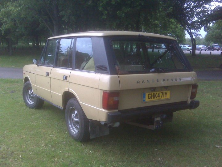 Restore Pistonheads Range Rover Classic - This image features a vintage Land Rover SUV parked on lush green grass in what appears to be a park or a similar outdoor setting. The car is painted in a beige color, and it has a distinctive black soft top roof that provides a contrasting element to the vehicle. The rear lights and a black bumper are also visible, adding to the overall robust and durable design of the Land Rover. The registration plate of the vehicle reads "GHK 47Y." The surroundings suggest a peaceful and serene environment, with trees and other foliage in the background.