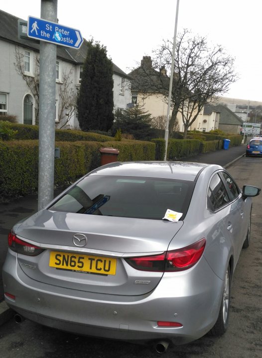 Lets play the Photo Location Game! Vol 3 - Page 48 - General Gassing - PistonHeads - The image shows a gray four-door sedan parked on the side of a street, next to a street sign. The car has a European-style license plate with black lettering on a yellow background. It's a clear day with a blue sky visible in the background. The car's side mirrors and the visible portion of the passenger side window reflect the surroundings, indicating that the photo was taken during the day.