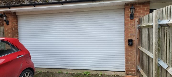 Volvo XC40 Recharge vs Skoda Enyaq - Page 5 - EV and Alternative Fuels - PistonHeads UK - The image shows a garage door with a wavy design, installed on the front of a house. It appears to be made of metal and has a modern, smooth finish. The color of the door contrasts with the surrounding environment, making it stand out. There are no visible texts or distinguishing marks that provide additional context or information about the location or property. The garage door is closed, and there's an indication of a red car parked in front of it.