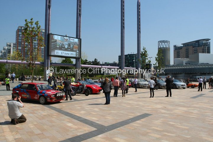 A group of people walking down a street - Pistonheads - The image showcases a city outdoor event, possibly a car show or filming. A large crowd of people is present with an array of cars on display, some cars appear to be in motion, and there is a group of photographers capturing the scene. In the backdrop, there are various buildings, including a grand, multistored edifice near the event, which adds an urban setting.