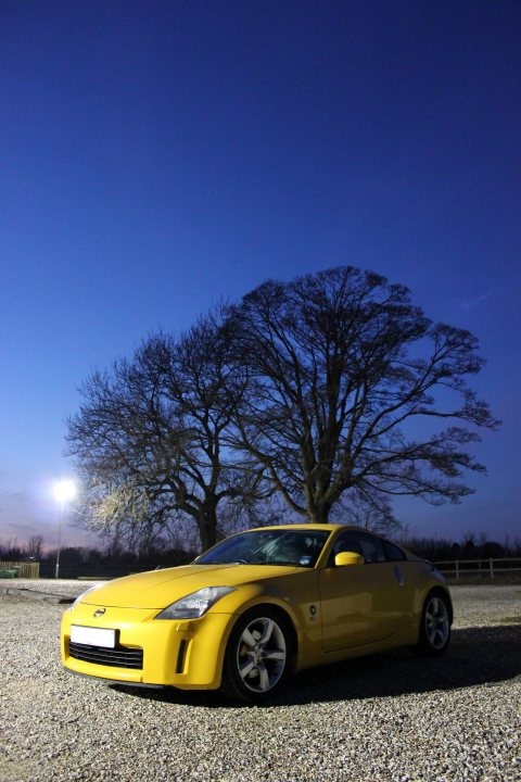 350Z GT4 - Ultra Yellow - Page 2 - Readers' Cars - PistonHeads - The image shows a yellow luxury sports car resting on a gravel surface. The car is parked, positioned facing towards the right side of the frame. A prominent feature is a large leafless tree beside the vehicle, against a deep blue sky. The lighting in the image suggests it may be late afternoon or evening, with the remaining light casting long shadows and highlighting the contours of the car and the rough texture of the gravel. There are no visible license plates or distinctive markings on the car. The style of the car and its color lay it open to judgments that can be made by observers about brand, model, or aspirational status.