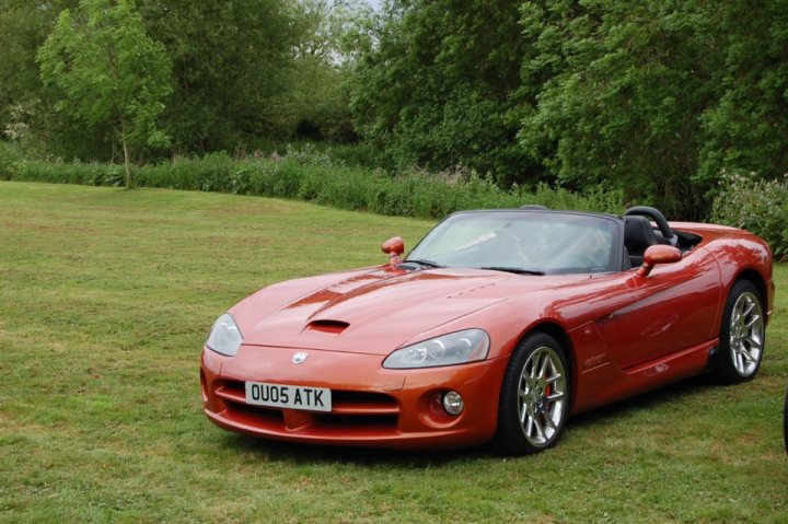 A red and white car parked in a field - Pistonheads - The image depicts a vibrant orange car parked on a lush green grassy area, possibly in a wooden field. The vehicle appears to be a convertible sports car, with the top down, revealing an interior. There is a license plate visible on the back of the car. The surrounding environment is verdant, with trees lining the perimeter of the image.