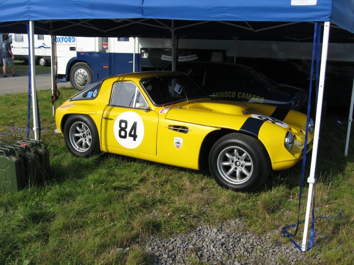 Early TVR Pictures - Page 126 - Classics - PistonHeads - The image showcases a vibrant yellow Morgan sports car, a classic model known for its distinctive design and engine, beautifully parked under a blue pop-up tent at a vintage auto show. This classic sports car is adorned with the number 84 and a striped hood, adding to its visual appeal. It's parked on a patch of grass amidst other classic cars, creating a setting that embraces automotive history. The scene is filled with a sense of nostalgia and appreciation for the engineering marvels of the past.