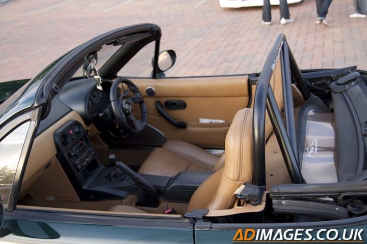 Buying an mx-5: What do i need to know? - Page 3 - Mazda MX5/Eunos/Miata - PistonHeads - The image depicts a convertible car interior with the top down, exposing the passenger seats. The vehicle has a beige or light tan interior, with the driver's seat visible. The steering wheel is on the left side, indicating that this is likely a UK-specified vehicle. A few smaller items are also visible, such as a rail and some other unidentifiable parts, possibly from the car's components. The setting appears to be on a brick pavement, and human figures can be seen in the background, but they are not the main focus of the image.