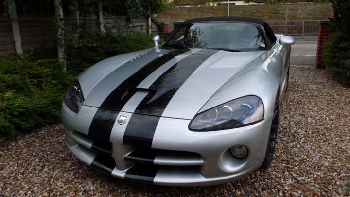 Pistonheads - The image shows a sports car parked on a gravel driveway. The car has a distinctive black and white racing stripe on the hood. The vehicle's design suggests high performance capabilities and it appears to be in a good condition. In the background, there is a well-manicured garden with trees and a fence, which are likely part of a residential property. The sky is visible with sunlight, indicating it's daytime. There are no visible texts or logos on the car.