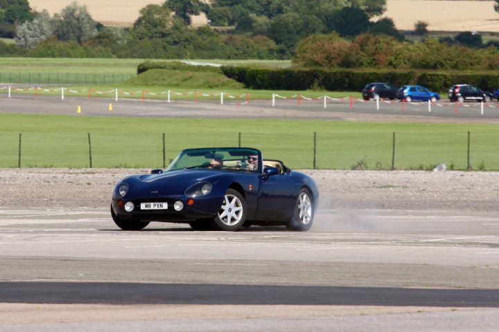 Sideways Griff Action at North Weald - Page 1 - Griffith - PistonHeads - The image captures a dynamic scene on a race track. A blue race car, sleek and low, is the center of attention as it occupies the center of the frame. The car is in motion, moving to the right of the image, creating a sense of speed and intensity. The track itself is bounded by a fence and lush green grass, providing a stark contrast to the hard lines and curves of the vehicle. The perspective of the photo is from behind, making the car the undeniable star of this track-side snapshot.