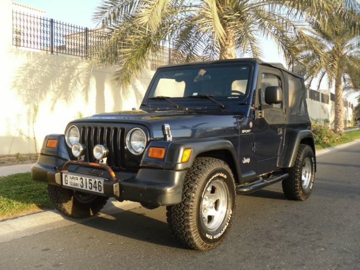 Pistonheads - This image shows a black off-road Jeep vehicle parked on the side of a street with palm trees in the background. The vehicle is equipped with furthest blue trim and has visible tinted windows and alloy wheels. It appears to be a sunny day with ample natural light washing over the scene. The Jeep has a rugged look, suggesting it is a utility or recreational vehicle intended for off-road use. The license plate is clearly visible on the front of the vehicle, indicating it in a region where such writing styles are required. The surroundings seem to be in an urban or suburban area.