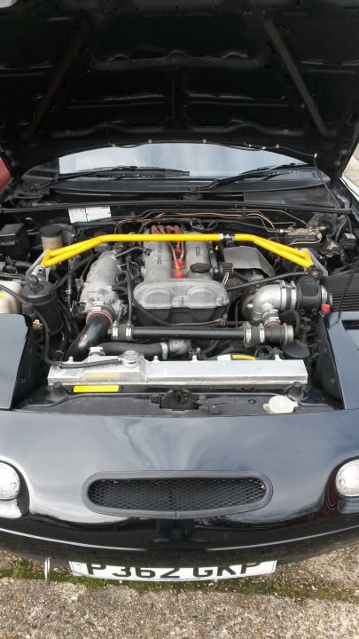 Who's the lucky one............... - Page 1 - Mazda MX5/Eunos/Miata - PistonHeads - The image shows the open hood of a sports car. Underneath the black hood, the visible engine components include a prominent yellow air duct, the bonnet, and various mechanical parts. The surrounding area appears to be a dirt or gravel surface, suggesting the car might be parked outdoors. The film-like appearance of the surface could indicate recent rain or dew, reflecting the light subtly. The photo is taken from a low angle, emphasizing the car's hood and some of the underside components.