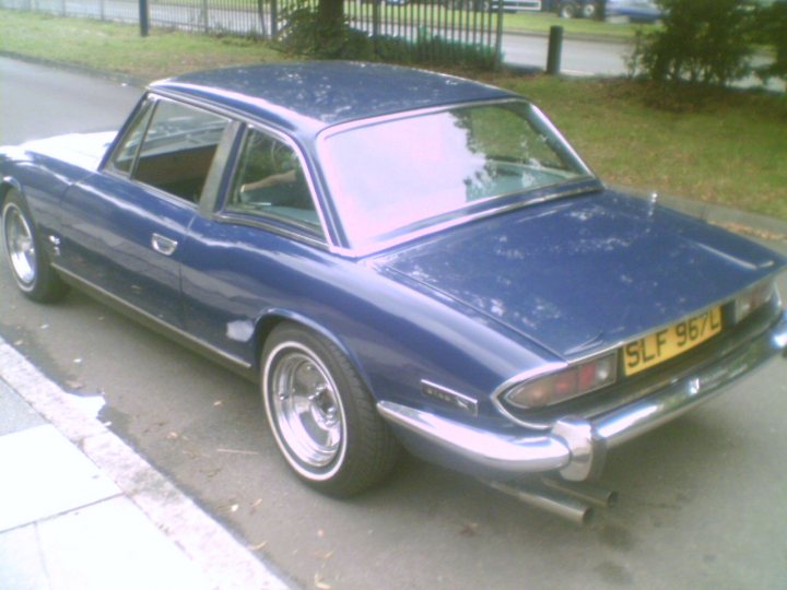 The image shows a classic two-door coupe parked on a roadside. The car is painted in a vibrant shade of blue and has chrome accents and a reflective roof. Its license plate reads "SLF 961". The car features a separate rear window, a distinctive feature for many classic vehicles. In the background, a portion of a fence is visible, suggesting the proximity of a property. The overall scene suggests a sunny day, given the visible shadows and the outdoor setting.