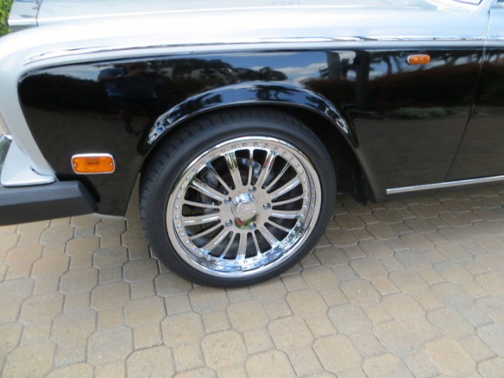 Silver Shadow Alloys... - Page 1 - Rolls Royce & Bentley - PistonHeads - The image displays the rear fender and wheel of a black car parked on a cobblestone surface. The vehicle appears to be a classic or modified model, indicated by the large, chrome-rimmed wheel and the polished finish of the fender and luggage compartment. The background is out of focus, which puts the emphasis on the car's distinctive features. The perspective suggests the photo was taken in an unobstructed area, as nothing else seems to be hindering a clear view of the car's details.