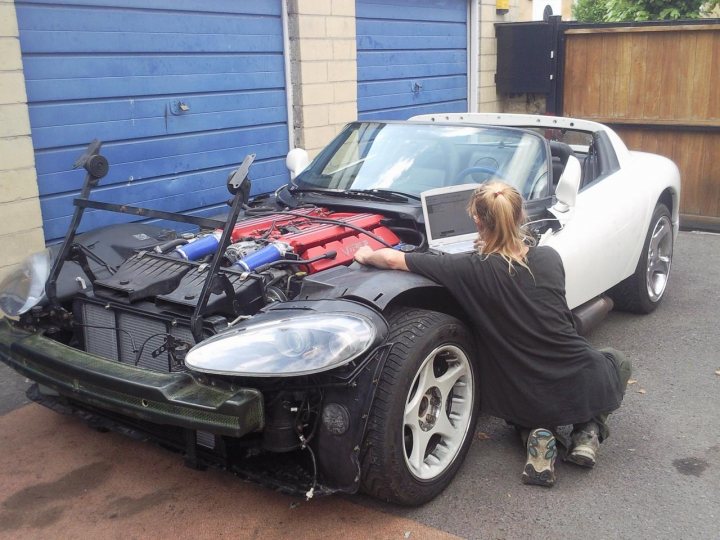 Rebuild Viper Pistonheads - A woman with blonde hair leans on the fender of a sports car under the hood, seemingly inspecting it. Inside the car, an open laptop is visible. The vehicle itself is a sleek, white sports car with a black hood that is raised. It is parked next to a fence and a garage.