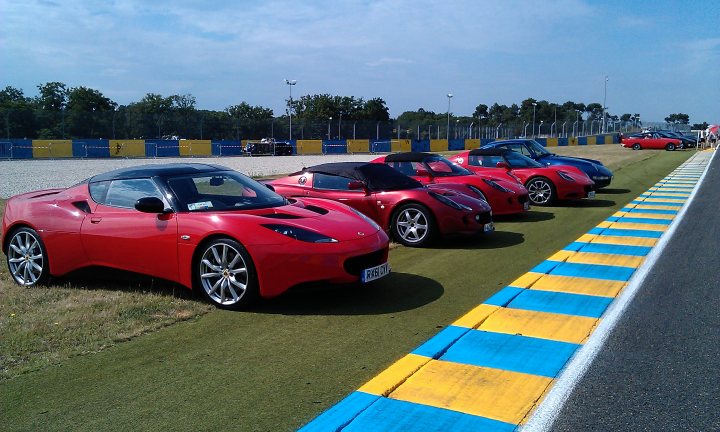 A red car is parked in a parking lot - Pistonheads - The image presents a serene scene on a race track. It features a line of six sports cars, five of which are sleek red sports cars, lined up on a track with a yellow and blue striped run-off area. These cars are parked closely together, capturing the essence of a race track. In the background, beyond the red sports cars, there are a couple of blue race cars. Further in the distance, you can spot a handful of people standing, possibly observers or staff. This setting suggests an organized event or practice session perhaps.