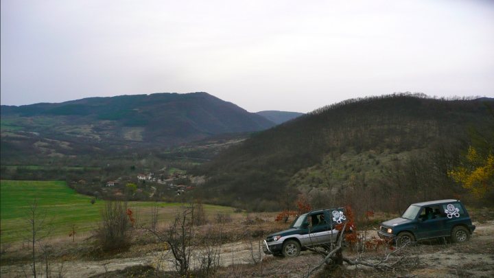 background car pics :) - Page 15 - Readers' Cars - PistonHeads - The image portrays a rugged outdoor scene with two SUVs parked on a dirt road in the foreground. The road cuts through an area with sparse vegetation, and beyond it, there's a mountainous landscape with rolling hills on the horizon under an overcast sky.