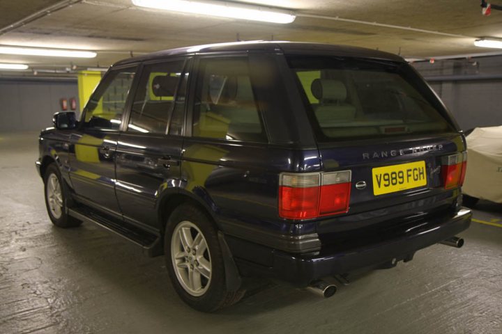 Range Rover P38 Ownership/Mpg? - Page 1 - Land Rover - PistonHeads - The image showcases a large, dark-colored Range Rover parked in a spacious garage area. The vehicle is positioned facing the camera, revealing its license plate which reads "Y99 FGH". The interior of the garage is mostly grey, with various yellow-toned strips and lights that provide illumination. A white vehicle, possibly a car, is partially visible in the background, suggesting that the garage is in use. There is a fire extinguisher on the left side of the image, indicating the safety measures in place within this facility.