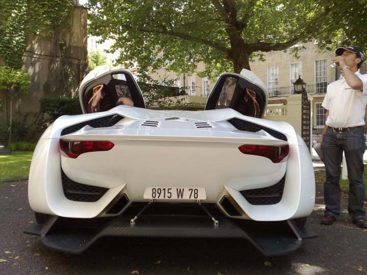 Supercars Rarities Pistonheads Spotted - The image captures a camouflaged white supercar parked in what appears to be a gated residential area, possibly a driveway. A person is standing beside the car, holding a drink bottle. The individual is taking a casual stance, with one hand in a pocket, and seems to be examining the sports car. The green environment suggests that the setting is outdoors, likely during a sunny day. The car is not visible in its entirety, but the unique rear design with a straight-cut tail section and LED taillights is distinct, indicative of the car's high-end and possibly sports-oriented nature.