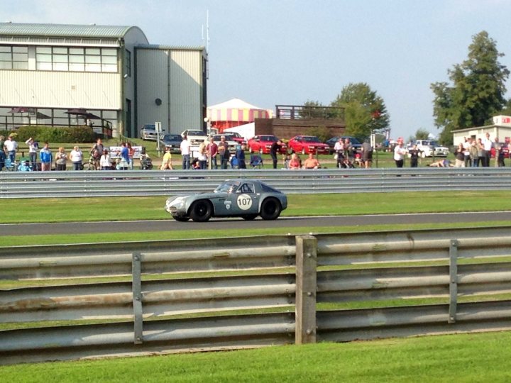 Early TVR Pictures - Page 126 - Classics - PistonHeads - The image is a photograph depicting a vintage car race. In the foreground, a classic racing car with distinctive post-war styling, including the air-intake scoop on its side and large round headlights, is driving on the race track, with the number 107 prominently visible on its side. The car is passing between protective barriers, typical of racing circuits, indicating that the car is participating in competitive racing. Behind the car, a crowd of onlookers watches the event, with some individuals taking photographs and videos using their handheld devices. Spectators are scattered around a grassy area that borders the race track, while cars are parked in a lot nearby. The background includes expansive grass fields, a large building with a distinct architectural design featuring a prominent circular element, and what appears to be a flagpole, all suggesting that this might be a major racing event on a clear day, possibly at dusk or close to sunset. The atmosphere is one of excitement and anticipation, common at motorsport events.