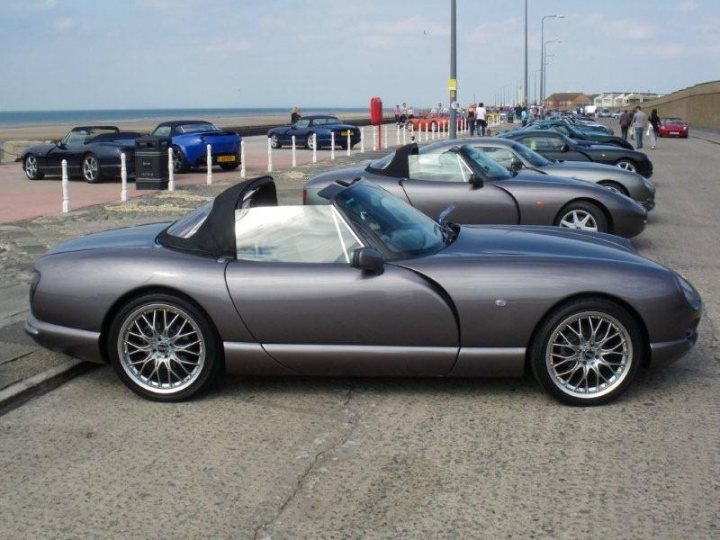 Chimaeras Aftermarket Alloys Pistonheads - The image shows a row of convertible sports cars parked parallel to each other along the edge of a pavement. The cars are sleek, with their convertible tops retracted, revealing their interiors. The parking is situated next to a beach with the sea visible in the background. A few individuals can be seen in the distance, adding to the relaxed, leisurely atmosphere of the scene.