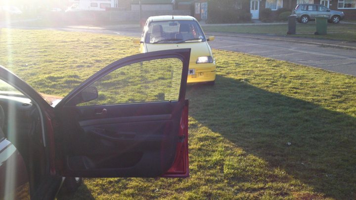 Driveway Diaries - Page 3 - East Anglia - PistonHeads - This is a photograph taken during the day, showing an open car door from a vehicle parked facing the rear of a second vehicle, which appears to be a small, yellow motorcycle or moped. The sunlight is casting a bright reflection and strong shadow across the image, suggesting that the photo was taken on a sunny day with the sun situated high in the sky. The camera angle is upward, capturing the perspective of the viewer looking from the open car door towards the front of the moped. There are residential houses and a road in the background with no people visible.