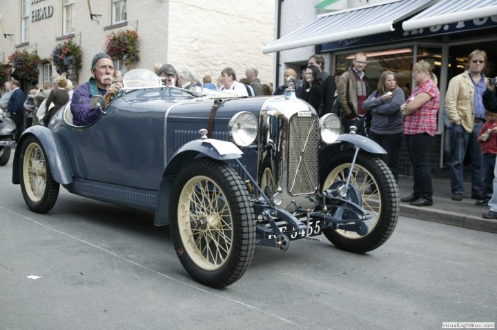 Classic tractors - Page 3 - Classic Cars and Yesterday's Heroes - PistonHeads - The image depicts a vintage car being driven on the street. The focal point of the image is the blue vehicle, which appears to be an antique with yellow wheels and a distinct radiator grill. Inside the car, there's a man driving, and another person is visible in the front passenger seat. The car is traveling down a road, and the passengers seem to be enjoying the ride. In the background, there's a crowd of people standing on the sidewalk, observing the car and possibly taking pictures. The street is lined with buildings, and the atmosphere appears to be casual and relaxed.