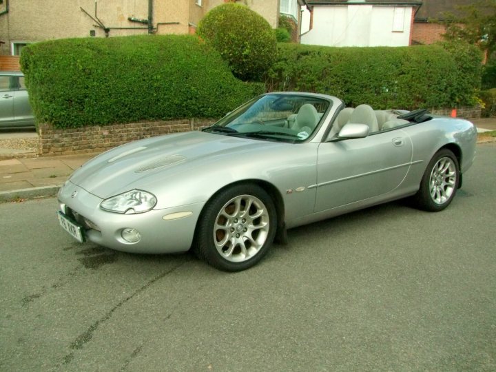 the new wheels !!!! - Page 1 - South West - PistonHeads - The image shows a sleek, silver-colored sports car parked on the street in front of a single-story house. The car, with its convertible top down, is the central focus of the photo. Its polished surface reflects the suburban residential setting. The house behind the car has a well-maintained lawn with a variety of bushes and trimmed hedges adding to the suburban aesthetic. The road on which the car is parked is empty, providing an uncluttered backdrop to the vehicle. This scene suggests the quieter moments of suburban living.