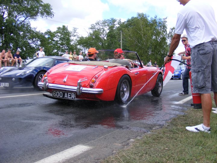 Pistonheads Classics - In the image, a vintage red sports car is participating in a parade, characterized by its black trimmings, chrome details, and the license plate 600 AER. The car is being sprayed with water from the side, likely for a cleaning or cooling purpose. People are standing on the roadside watching the action, with some comfortably seated on the grass. A person can be observed directing traffic with a hose connected to the car, ensuring safety and order during this special event. The atmosphere suggests a community gathering, possibly a car show or festival, under a sunny sky.