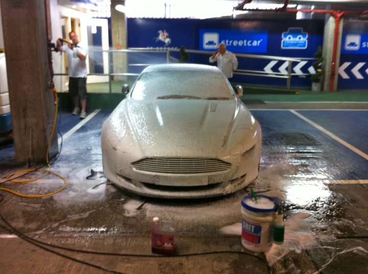 Pistonheads - In the image, a gray sports car is undergoing a car wash in a brightly lit station. The car is parked on a wet floor with a visible foam suction liquid being used during the wash process. Two workers, one using a pressure gun and another holding a bucket, are meticulously cleaning the car. The cars 'Streetcar' branding is evident, suggesting the location or service offered. A sticker on the windshield indicates that a car wash solvent has been applied to the glazing.